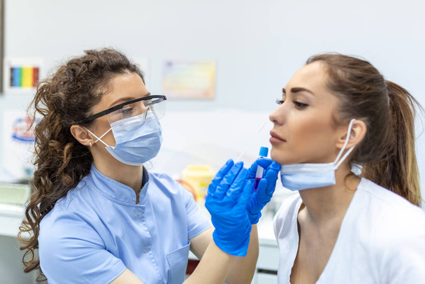 マスクを着用している女性患者は、 Covid 19のpCR検査のために鼻からサンプルを採取しています。保護具付きの医療従事者は女性にコロナウイルスの波を実行します。鼻スワブfor Covid-19. - 写真・画像