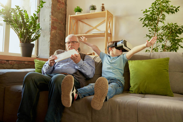 Modern gadgets. Portrait of happy senior man and little boy, grandfather and grandson using VR glasses sitting on sofa at home and laughing. Hobbies, leisure activities, family and emotions - Fotó, kép