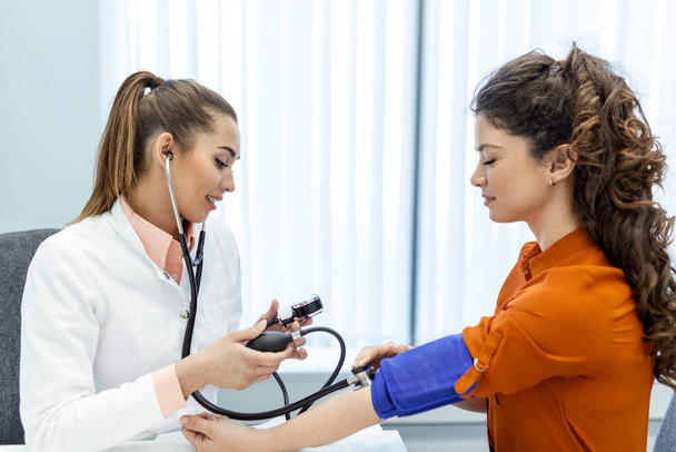 A medir a pressão arterial. Jovem mulher tem uma visita com médico feminino na clínica moderna. A medir a pressão arterial. Manómetro. Hipertensão e frequência cardíaca elevada - Foto, Imagem