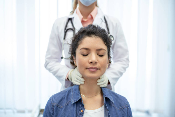 Ενδοκρινολόγος εξετάζει το λαιμό μιας νεαρής γυναίκας στην κλινική. Γυναίκες με εξέταση θυρεοειδούς αδένα. Ενδοκρινολογία, ορμόνες και θεραπεία. Φλεγμονή του πονόλαιμου - Φωτογραφία, εικόνα