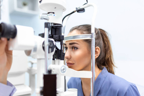 Attente optometrist onderzoekt vrouwelijke patiënt op spleetlamp in oogheelkundige kliniek. Jonge mooie vrouw is gediagnosticeerd met oogdruk op speciale oogheelkundige apparatuur. - Foto, afbeelding