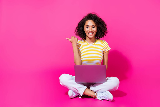 Teljes testméret fotó fiatal ülő lány ujját mutató üres tér használata számítógép izgatott pozitív elszigetelt rózsaszín színű háttér. - Fotó, kép