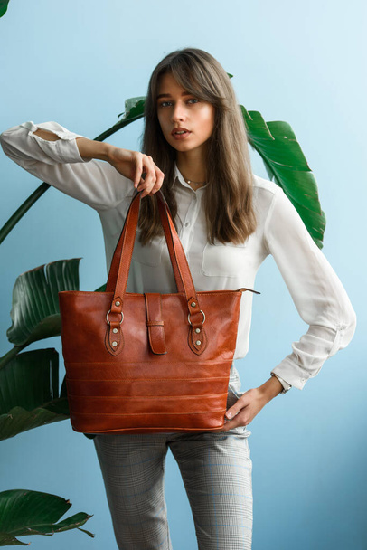 close-up photo of orange leather bag in a womans hands. indoor photo. beautiful slender girl in gray plaid pants and a white blouse posing with a handbag - Photo, Image