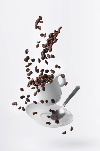 granos de café tostados derramados de una taza flotante y una cucharadita aislada sobre fondo blanco - Foto, imagen