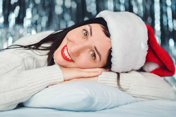Close up portrait of beautiful smiling brunette young woman with red lipstick, white sweater, Santa Claus hat lying on bed at home, happy Christmas mood. Cheerful lady with New Year sparkle lights - Photo, Image