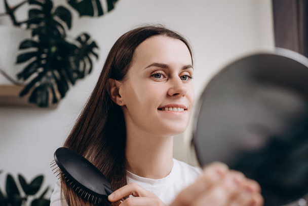 Cheerful attractive young lady sit on couch at home, combing her beautiful silky hair with wooden brush, looking at mirror and smiling. Cute female do morning beauty routineHair care products concept - Fotó, kép