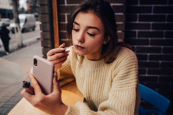 Smiling woman dressed warm sweater, hold phone gadget and making hurry makeup in street cafe, Beauty, lip liner lipstick - Photo, Image