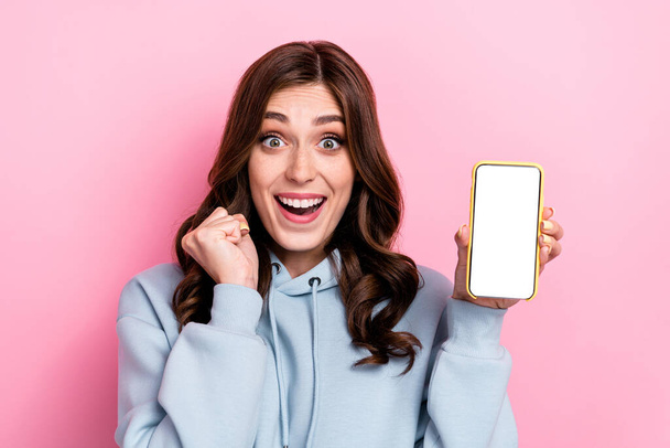 Portrait of excited crazy girl hold show empty space telephone display raise fist isolated on pink color background. - Foto, afbeelding