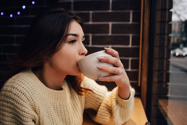 Lächelnde Frau trägt warmen Pullover, trinkt Marshmallow mit Milchkakao oder Latte. Heißes aromatisches Schokoladengetränk. Wärmendes Snack-Essen im Straßencafé. Neujahr Weihnachtsschmuck, Girlanden - Foto, Bild