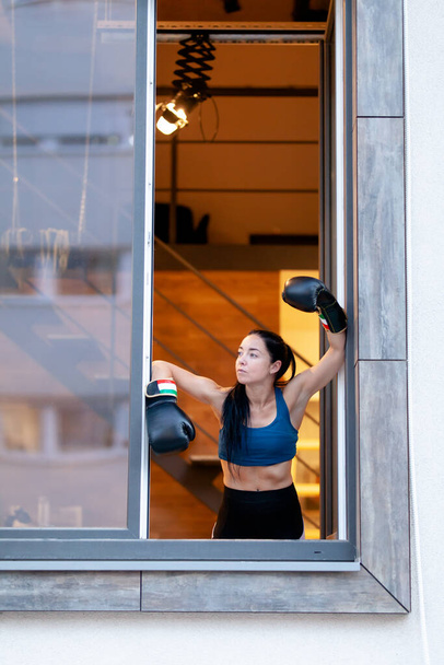 Fit girl with boxing gloves relaxing and looking out of window during break from exercise. - Foto, Imagem