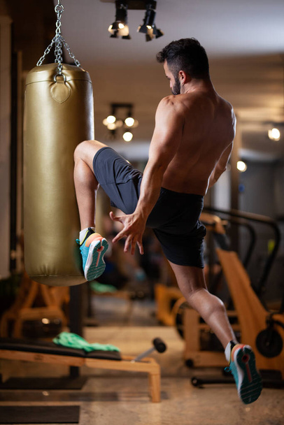 Muscular shirtless man jump hitting a punching bag with a knee in a fitness studio. - Foto, immagini