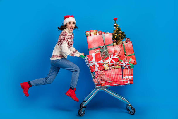 Photo of cool impressed lady wear print pullover santa headwear jumping buying presents shopping tray isolated blue color background. - Φωτογραφία, εικόνα