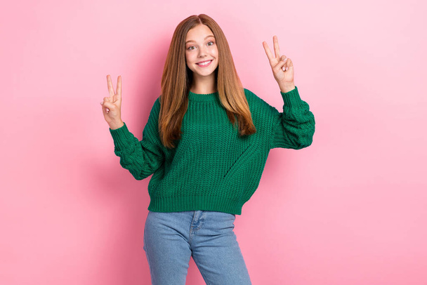 若い人気ブロガーの女の子の写真ポーズを示すv-sign肯定的な笑顔は、スタイリッシュな衣装を身に着けています緑のジャンパーデニムはパステルピンクの色の背景に隔離. - 写真・画像