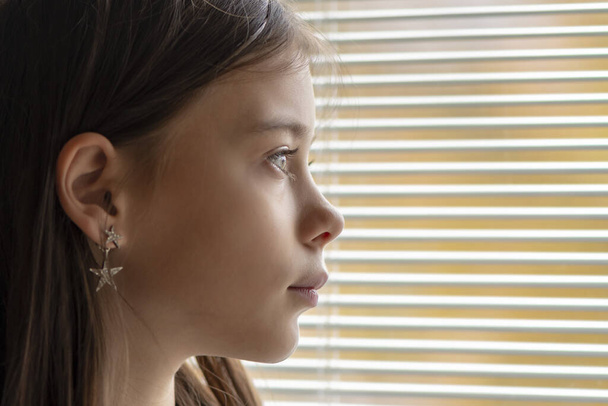 Een 11-jarig meisje kijkt door de verlaagde jaloezieën van het raam op straat, close-up, selectieve focus. Concept: wachten op ouders, weeshuis, alimentatie. - Foto, afbeelding