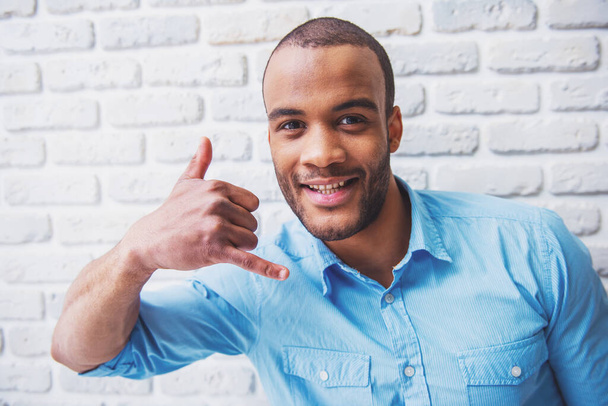 Portrait of handsome Afro American man showing hand phone, looking at camera and smiling, against white brick wall - Photo, Image