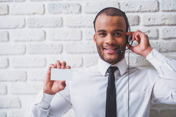 Csinos afro-amerikai üzletember, klasszikus ruhák és audio headset mutatja kártya, nézte a kamerát, és mosolyogva, szemben a fehér téglafal - Fotó, kép