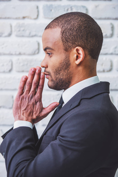 Vue latérale d'un bel homme d'affaires afro-américain en costume classique, les mains jointes comme pour prier, contre un mur de briques blanches
 - Photo, image