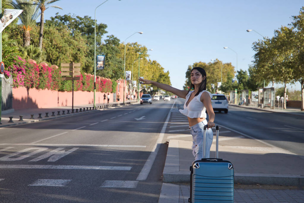 Donna sudamericana, giovane e bella, bruna, con camicia bianca, jeans e una valigia, che chiede un taxi. Concetto viaggi, trasporti, vacanze, bagagli, taxi. - Foto, immagini