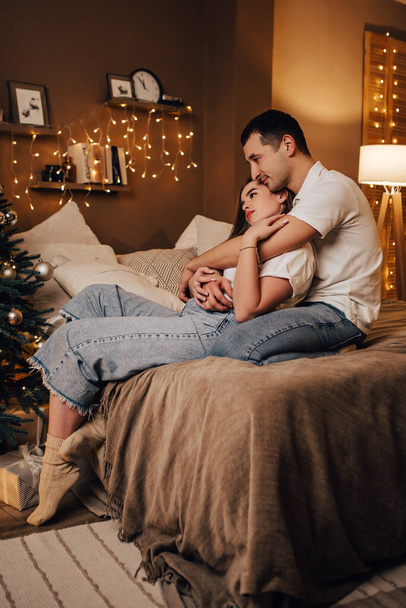 Vertical image lovely young romantic couple lying on bed garlands kissing hugging home cosy interior atmosphere New Year Christmas tree decorations holiday party celebrating concept winter evening  - Photo, image