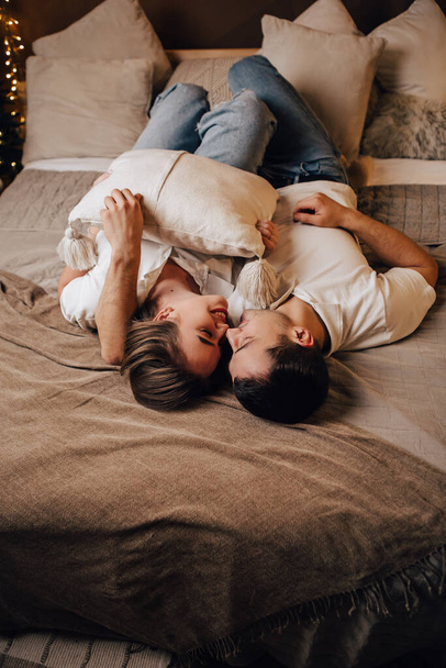 Vertical image lovely young romantic couple lying on bed garlands kissing hugging home cosy interior atmosphere New Year Christmas tree decorations holiday party celebrating concept winter evening  - Photo, Image