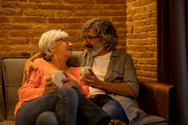 Старшая пара с седыми волосами обнимается и смотрит друг на друга, выпивая горячий напиток в кафе - Фото, изображение