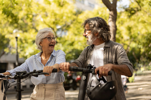 Close up feliz casal de cabelos grisalhos seniores em empurrar scooter equitação em áreas verdes - Foto, Imagem