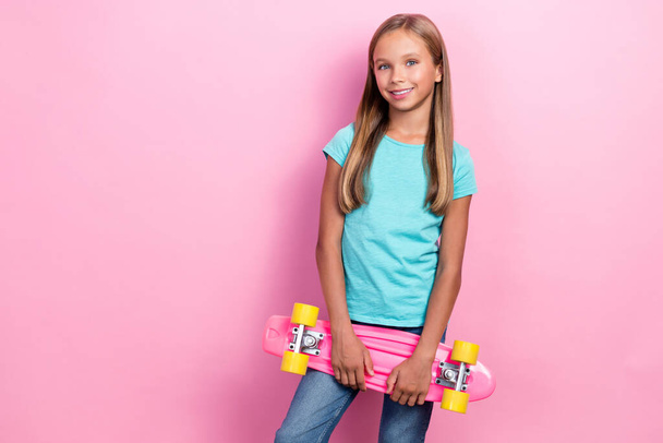 Photo of sweet shiny small kid wear turquoise t-shirt holding skateboard empty space isolated pink color background. - Foto, imagen