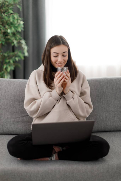 Happy attractive student enjoying warm drink while using portable laptop. Charming young female in beige hoodie spending leisure time with gadgets at home. - Foto, afbeelding