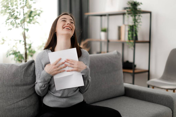 Überglückliche Frau mit braunen Haaren freut sich über Brief von der Universität. Fröhliche junge Studentin liest Dokument über Bildung, während sie zu Hause auf dem Sofa sitzt. - Foto, Bild