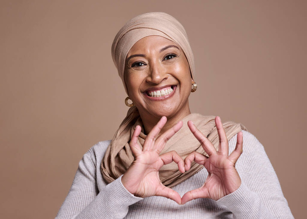 Islam woman, heart and sign with smile, happy and peace against brown studio background. Muslim lady, mature female and hand gesture for happiness, love and commitment for humanity and positive - Valokuva, kuva