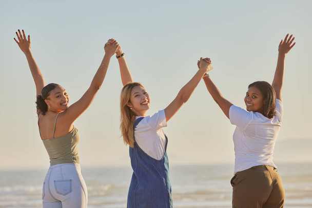 Happy, girl friends and holding hands at a beach with women on a holiday, freedom and travel. Portrait of people with arms raised to show excited adventure by the ocean and sea together by water. - Фото, изображение