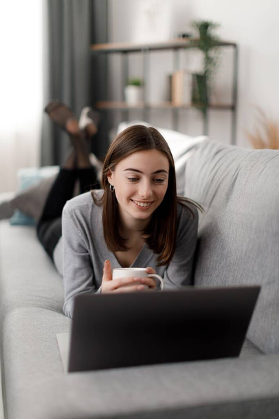 Šťastná mladá žena pomocí bezdrátového notebooku pro sledování filmů, zatímco leží doma na pohovce. Příjemná mladá dáma drží šálek horkého nápoje v rukou a těší relaxaci. - Fotografie, Obrázek