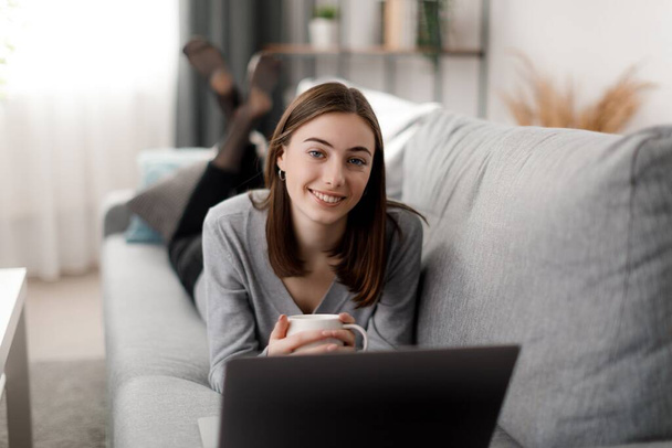 Attraktive junge Frau mit braunen Haaren mit drahtlosem Laptop und Teetrinken auf der gemütlichen Couch. Häuslicher Lebensstil und Technologiekonzept. - Foto, Bild