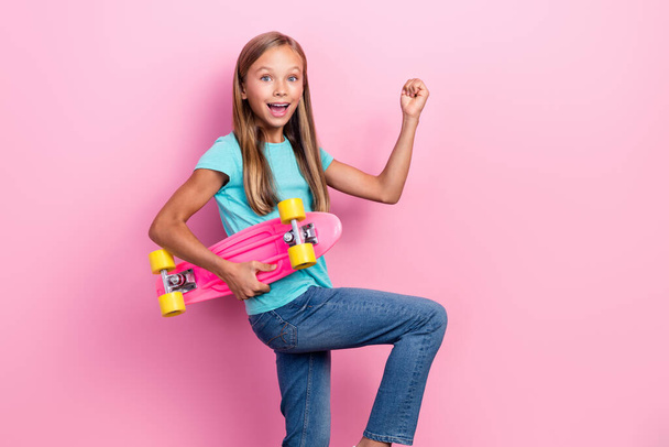 Photo of adorable lucky small kid wear turquoise t-shirt riding skateboard rising fist isolated pink color background. - Foto, imagen