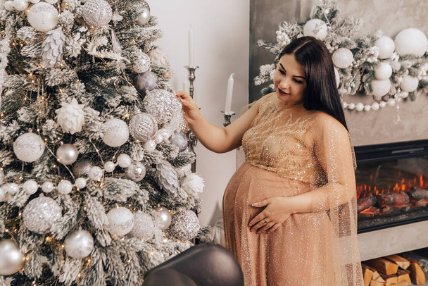 Woman with round belly with baby kid inside decorating Christmas Tree, celebrating New Year holiday. Pregnant lady waiting for infant. Expecting a newborn child - Foto, Imagem
