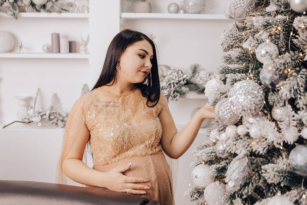 Karnında bebek olan bir kadın Noel Ağacı 'nı süslüyor, yeni yılı kutluyor. Bebek bekleyen hamile bir kadın. Yeni doğmuş bir çocuk bekliyorum. - Fotoğraf, Görsel