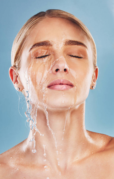 Water, skincare and face of a woman with a splash for cleaning, treatment and care against a blue studio background. Wellness, health and girl model with luxury facial dermatology to relax for beauty. - Photo, Image