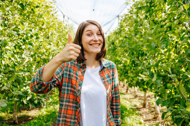 Thumb up with one hand wide smile young woman farmer looking at camera. Copy space in an apple orchard. She is wearing a plaid shirt and a white t shirt. Front view. - Fotoğraf, Görsel