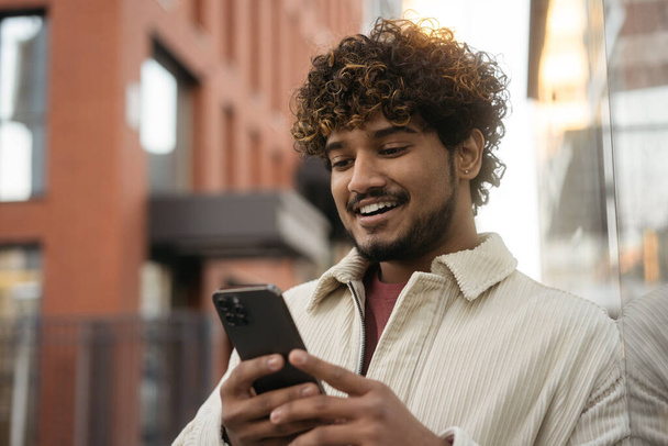 Mosolygós göndör hajú indiai férfi használ mobil app vásárlás online, válasszon valamit a honlapon. Boldog ázsiai hipszter srác játszik mobil játék áll a város utcáján. Modern technológiai koncepció  - Fotó, kép