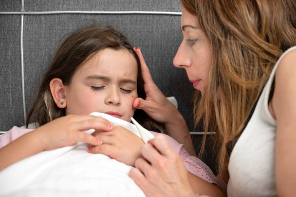 Matka měří teplotu své nemocné dcery. Nemocné dítě s vysokou horečkou ležící v posteli a matka držící teploměr. Kvalitní fotografie - Fotografie, Obrázek