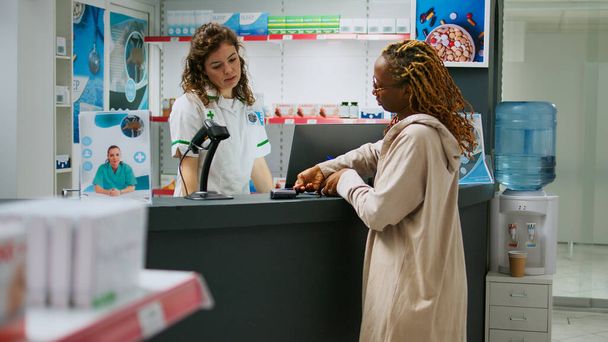 African american client buying pharmaceutical products and paying with credit card at pharmacy counter. Using contactless payment to buy medicaments and healthcare supplements at drugstore. - Photo, Image