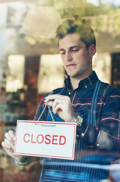 Time to close up for the day. Closeup shot of a young man hanging up a closed sign in a shop window - Photo, Image