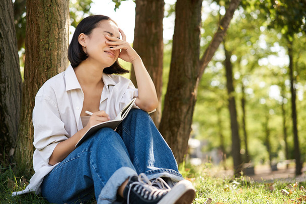 Szomorú lány sír, miközben a naplójába ír, egyedül ül a fa alatt a parkban a jegyzetfüzetével, zokog és vigyorog.. - Fotó, kép
