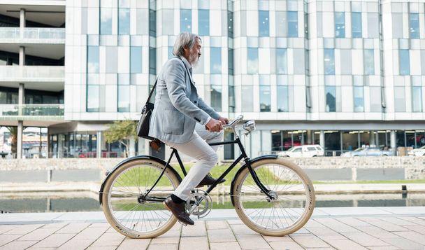 Travel, city and Asian businessman on bicycle, cycling to work in urban town for low carbon footprint. Sustainability, transport and senior man riding bike to workplace for eco friendly commute. - Photo, Image