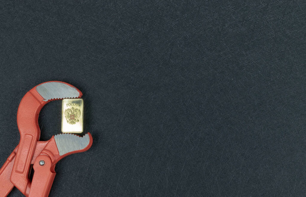 РОССИЯ монета золотая в челюстях трубного ключа. Инфляция на металлической пластинке РОССИЯ война Украина. концепт символ - Фото, изображение