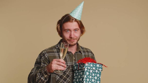 Joyful happy young man with gift box dancing, celebrating champagne drinking glass. Birthday surprise disco party. Young guy boy satisfied with best present bonus isolated on beige studio background - Photo, Image