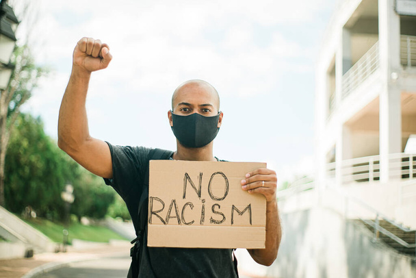 Dark skinned young man in a black mask holds a poster with the inscription No Racism in one hand, while the other hand is clenched into a fist above his head - Photo, image