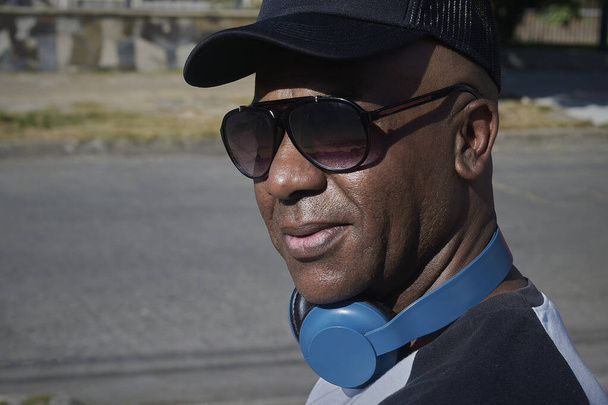 Πορτρέτο ενός ενήλικα Αφροαμερικανού με γυαλιά ηλίου, ακουστικά και μαύρο καπέλο. - Φωτογραφία, εικόνα