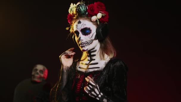 Kobieta śmierci słuchająca muzyki na słuchawkach, nosząca makijaż czaszki i kostium Mikołaja Muerte w dniu śmierci. Ciesząc się zabawną piosenką na słuchawkach audio, świętując meksykańską tradycję. Strzał ręczny. - Materiał filmowy, wideo