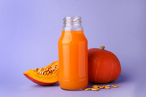 Tasty pumpkin juice in glass bottle, whole and cut pumpkins on lavender color background - Valokuva, kuva